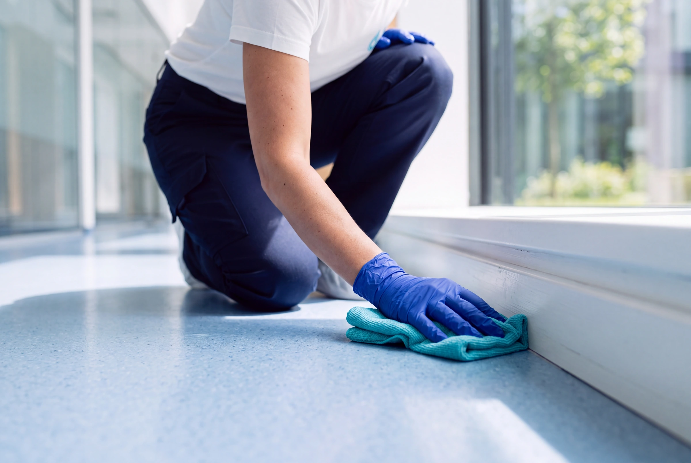 A blue-gloved hand cleans the floor beside the skirting with a turquoise cloth.